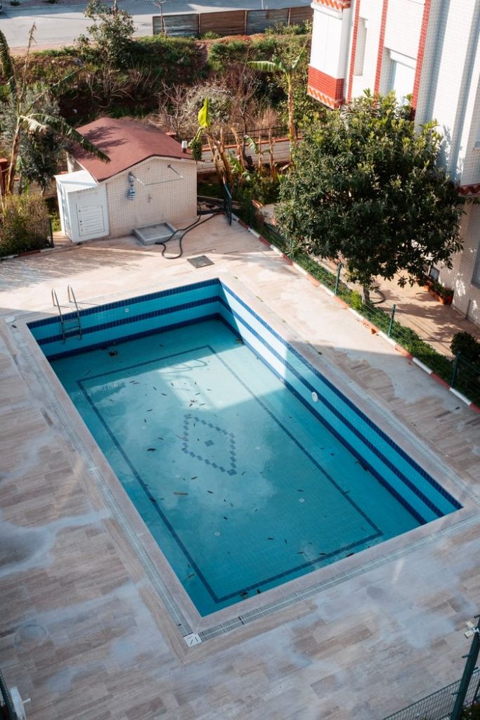 An empty swimming pool in a sunlit garden in Antalya, Türkiye, surrounded by lush greenery.